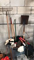 Photo showing several garden tools including rake, shovel, tree trimmer, bolt cutters, hand pruners, rubber boots, and gloves arranged around a trashcan.