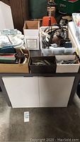 Photo of metal cabinet with white doors on wheels, with multiple boxes of handyman parts on top including metal hinges, electrical cords, and assorted hardware.