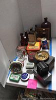Photo showing collection of vintage bottles, tins, clock, glass mug, and small collectibles arranged on table.