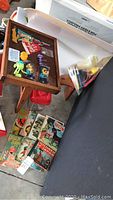 Small vintage wooden peg desk with various small figurines and keychains on top, next to a stack of vintage comic and paperback books on the floor.