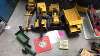 Three yellow Tonka metal construction vehicles, one green metal John Deere tractor, and two books on floor.