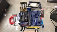 Top view of scattered tools showing Stanley handsaw, red C-clamps, screwdrivers, socket set in blue case, hacksaw, and other items