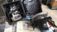 Craftsman router and circular saw in their black carrying cases, with manuals visible beneath them on the floor.