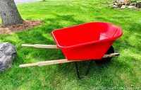 Side view of the red wheelbarrow showing the deep red plastic tub with wooden handles and one black tire on a metal frame on grass outdoors.