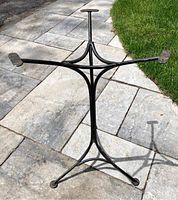 Photo of vintage cast iron table base standing outdoors on stone pavers and grass, showing full structure and three legs.