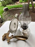 Full view of antique oil lamp with glass chimney, font, and metal reflector attached to ornate cast iron wall mount bracket on white surface outdoors.
