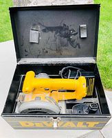 Top-down view of yellow DeWalt cordless circular saw and charger inside the black metal DeWalt case, showing saw handle, blade guard, charger with label, and lure fastening straps on case lid.