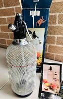Vintage Syphon soda seltzer bottle covered with a silver metal mesh sleeve, black and chrome dispenser top, under daylight against brick wall, original box and pamphlets visible.
