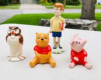 Front view of all four porcelain character figures from Winnie the Pooh by Beswick: Owl, Winnie the Pooh, Piglet, and Christopher Robin standing on white surface outdoors with background showing stone and grass.