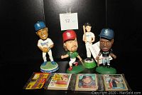 Photo showing four bobbleheads of baseball players from Dodgers, Cardinals, Mariners, and Blue Jays along with three individual vintage baseball cards and one sealed 1987 Topps 48-card pack.