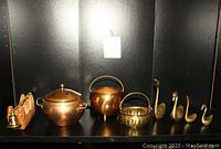 Photo showing copper pot with lid and handles, copper kettle, ribbed brass bowl with braided handle, four brass swan figurines, and brass candle extinguisher all arranged side by side against a dark background