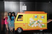 Three Barbie dolls standing next to a vintage yellow and orange Barbie Country Camper toy vehicle with floral graphics.