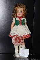 Full view of the Shirley Temple doll standing on a metal stand, wearing a green and red dress with a white apron and a Shirley Temple vintage pin attached to the dress.