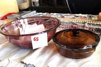Photo of the Pyrex purple glass bowl and Anchor Hocking brown glass covered casserole dish side by side on a surface, with edge of fabric and furniture visible.