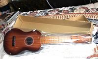 Ukulele with sunburst finish, sealed in plastic, lying next to open cardboard box on patterned surface.
