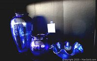 Three vintage blue glass decorative items on black shelf: large tall vase, medium rounded vase, and ribbon-edged bowl