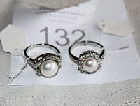 Two women's rings showing central pearls surrounded by clear rhinestones on silver-tone bands, placed on a white cloth with tags.