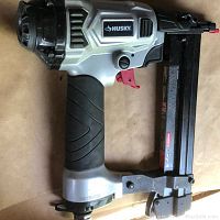 Side view of Husky pneumatic nailer showing the Husky logo, silver and black housing with ergonomic rubber grip and red trigger.