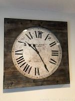 Large square wall clock with Roman numerals and black hands mounted on dark wood.