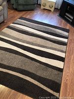 Full view of rectangular accent rug showing wave pattern in black, white and different grays on wooden floor near couch and furniture.