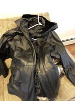 Photo showing front of black leather jacket with zip and button closure, jacket hung on a white hanger.