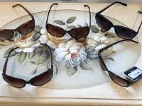 Five sunglasses laid out on a floral surface showing different frame styles and lens shapes.