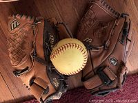 Lot with two brown softball/baseball gloves featuring black lacing, woven pattern, and one yellow softball on wooden floor.