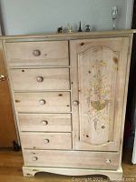 Front view of Vaughan of Virginia wooden armoire chest with floral painted door and five drawers.