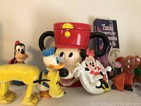 Photo of assorted Disney character figurines and large Mickey Mouse mug with ear handles including Donald Duck, Pluto, Goofy, Minnie Mouse, and more.