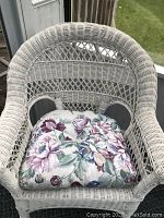 Front angled view of the white wicker chair showing woven backrest and floral cushion on seat.