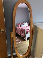 Front view of tall oval cheval floor mirror with oak frame standing on hardwood floor near a bedroom.