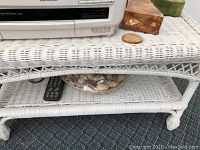 Close-up of the white wicker entertainment center lower shelf with seashell bowl, remote, and part of the stand legs with woven wicker texture.
