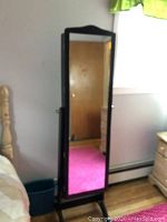 Full view of the tall black cheval standing mirror chest situated next to a bed and window.