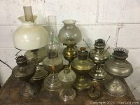 Front view of group of eight vintage and antique oil lamps on wooden surface with concrete wall background.