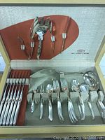 Open silverplate cutlery box showing eight place settings of knives, forks, spoons, plus additional large serving utensils including serving fork and ladle. Orange satin fabric lining in box lid. Polished stainless handles with a mid century modern design.