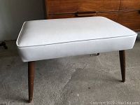 Photo of a rectangular white vinyl cushioned stool with four angled wooden legs showing scratches and wear.