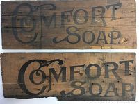 Photo showing two vintage wooden signs with 'COMFORT SOAP' painted in black on aged wood.