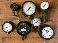Seven assorted pressure gauges laid out on wooden surface showing fronts of dials and connections.
