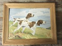 Full view of framed paint-by-number painting showing two brown and white dogs in grassy landscape under cloudy sky.