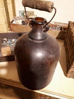 Photo of a large antique stoneware jug with dark brown glaze and wooden handle on metal frame