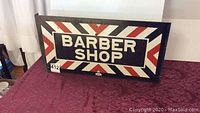 Front view of sign showing “Barber Shop” text and diagonal red and white stripes on black background.