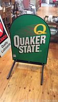 Front view of Quaker State sidewalk sign on wooden floor, clearly showing green background and logo.