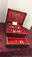 Full view of open wooden chest showing upper and lower compartments filled with silver plate cutlery items arranged in red velvet lining.