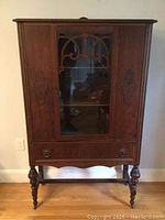 Front view of the antique high boy china cabinet showing the closed glass front door, drawer, and turned legs.
