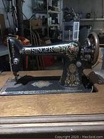 Front and side view of ornate black Singer 99 Red Eye sewing machine with gold and red decoration, set in original wood table with storage drawers.