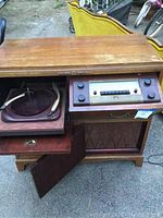 Photo of the stereo cabinet with the top open showing the turntable, tone arm, radio control panel with tuning dial and knobs, speaker grille below, and cabinet legs
