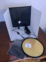 White pop-up light box setup with two LED lights inside, cords visible, circular gold/silver reflector and fabric backdrops stacked nearby on table.