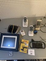 Wide shot showing all items on table: digital photo frame with remote, silver iPod Classic, two cordless phones, vintage Palm Pocket PC, black wireless modem, laser device in packaging.