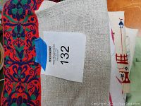 Photo showing edge of a tablecloth with hemstitched green fabric and part of a red and blue floral patterned runner underneath, featuring MaxSold lot number sticker.