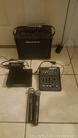Photo showing Blackstar amplifier, Alesis audio mixer, Innopow wireless microphone receiver, and two handheld microphones with cables on tile floor.